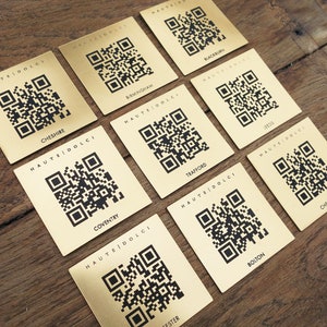 May include: A selection of gold-toned square cards featuring black QR codes and city names like "Cheshire" and "Birmingham". The cards are arranged on a wooden surface. Each card displays the text "HAUTEIDOLCI".