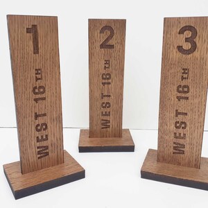 May include: Three wooden table number signs. Each sign is a rectangular block with a number etched at the top and the text "WEST 16TH" vertically down the centre. The numbers are 1, 2, and 3. The signs are brown with a dark brown base.