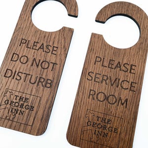 May include: Two wooden door hangers with a dark brown finish. One reads "PLEASE DO NOT DISTURB" and the other "PLEASE SERVICE ROOM." Both have "THE GEORGE INN" logo. The hangers have a curved top for hanging.