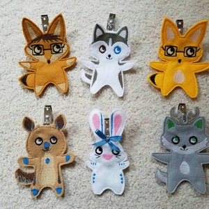 May include: Six felt animal plushies with embroidered details. The plushies are a fox wearing glasses, a husky, a fox, a bunny, a dog, and a wolf. Each plushie has a silver clip attached to the top.