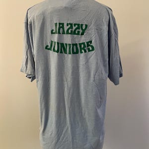 Może przedstawiać: Jasnoniebieska koszulka z nadrukiem "Jazzy Juniors" w kolorze zielonym na plecach.