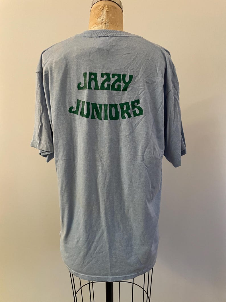 Może przedstawiać: Jasnoniebieska koszulka z nadrukiem "Jazzy Juniors" w kolorze zielonym na plecach.