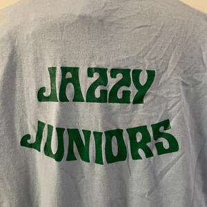 Może przedstawiać: Jasnoniebieska koszulka z nadrukiem "Jazzy Juniors" w kolorze zielonym.
