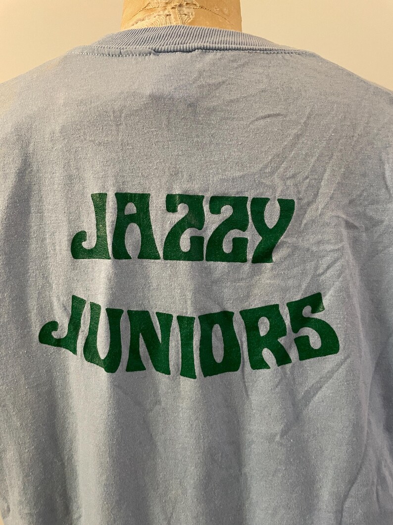 Może przedstawiać: Jasnoniebieska koszulka z nadrukiem "Jazzy Juniors" w kolorze zielonym.