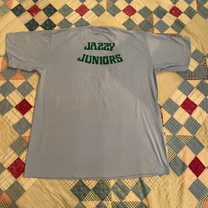 Może przedstawiać: Jasno niebieski t-shirt z napisem "Jazzy Juniors" w zielonych literach na plecach.