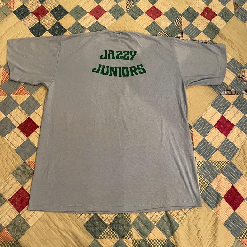 Może przedstawiać: Jasno niebieski t-shirt z napisem "Jazzy Juniors" w zielonych literach na plecach.