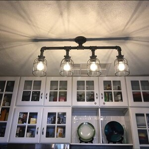 May include: A four-light industrial style ceiling fixture with caged glass shades. The fixture is made of black metal pipes and is mounted to a white ceiling. The fixture is hanging over a white kitchen cabinet with glass doors.