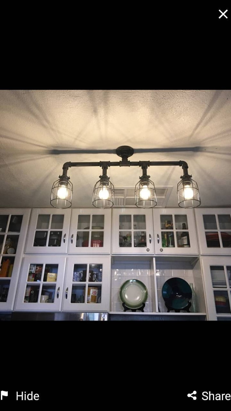 May include: A four-light industrial style ceiling fixture with caged glass shades. The fixture is made of black metal pipes and is mounted to a white ceiling. The fixture is hanging over a white kitchen cabinet with glass doors.