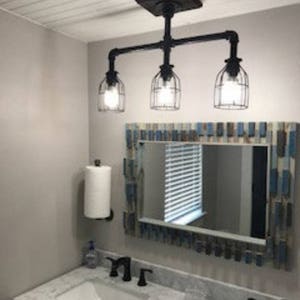 May include: A bathroom vanity with a white marble countertop and two sinks. There are black faucets and a black pipe light fixture with three caged light bulbs. A mirror with a blue and white frame hangs above the sinks.