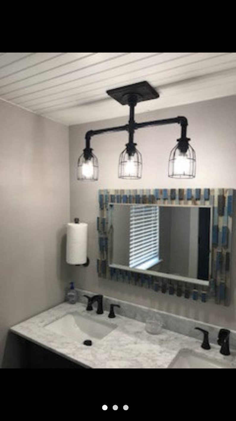 May include: A bathroom vanity with a white marble countertop and two sinks. There are black faucets and a black pipe light fixture with three caged light bulbs. A mirror with a blue and white frame hangs above the sinks.