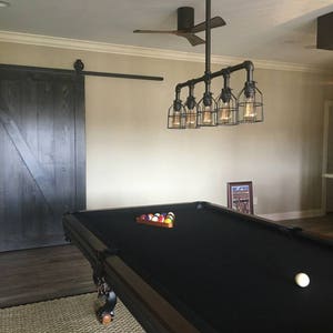 May include: A black pool table with a black felt surface and a set of billiard balls on the table. A white billiard ball is on the felt surface. A black ceiling fan with wooden blades is in the background. A black metal pipe light fixture with six caged light bulbs hangs above the pool table.