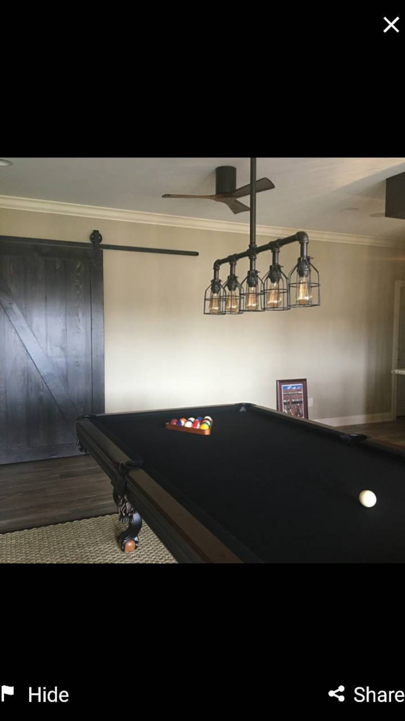 May include: A black pool table with a black felt surface and a set of billiard balls on the table. A white billiard ball is on the felt surface. A black ceiling fan with wooden blades is in the background. A black metal pipe light fixture with six caged light bulbs hangs above the pool table.