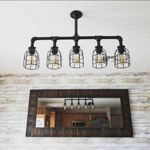 May include: A black industrial style pipe chandelier with five caged light bulbs. The chandelier is hanging from the ceiling and is reflected in a brown wooden framed mirror.