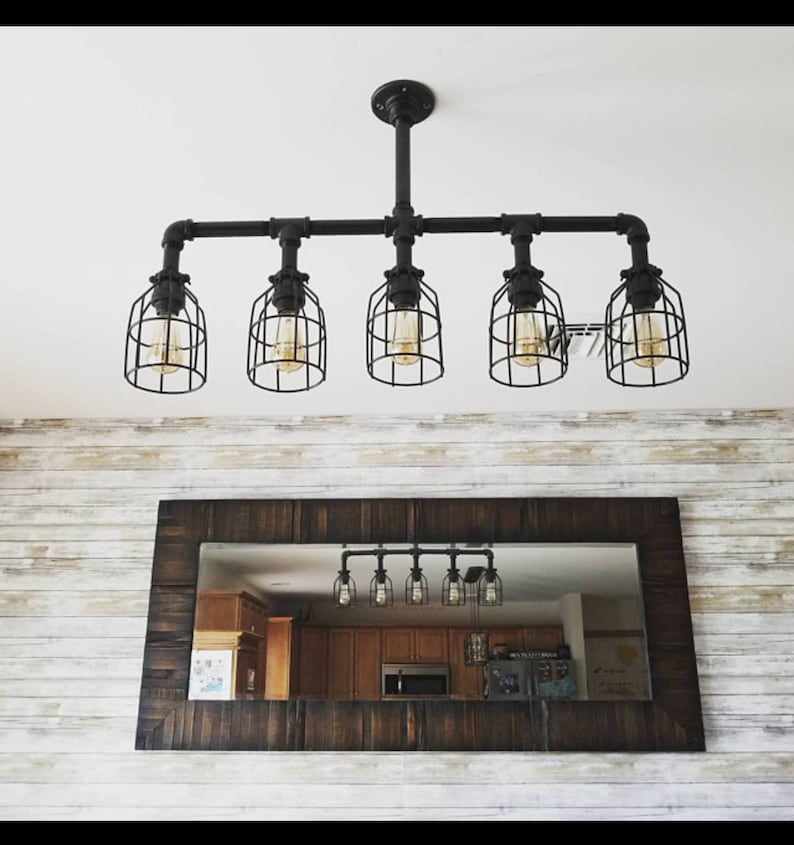 May include: A black industrial style pipe chandelier with five caged light bulbs. The chandelier is hanging from the ceiling and is reflected in a brown wooden framed mirror.
