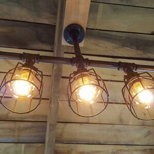 May include: Five caged light bulbs hang from a black metal pipe fixture. The lights are suspended from a wooden ceiling.