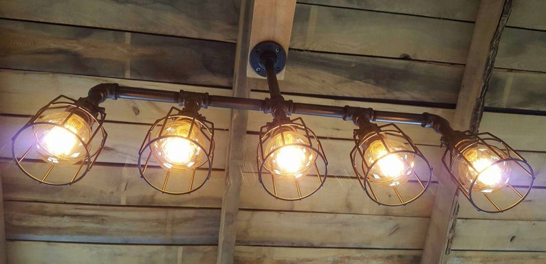 May include: Five caged light bulbs hang from a black metal pipe fixture. The lights are suspended from a wooden ceiling.