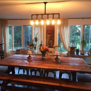 May include: A rustic wooden dining table with a long, rectangular top and a bench seat. The table is lit by a six-light industrial-style chandelier with exposed pipes and clear glass bulbs.
