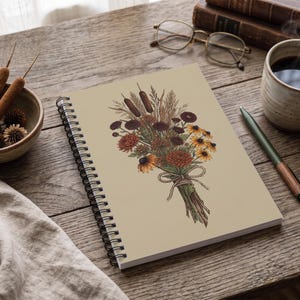 Può includere: Un taccuino a spirale con copertina color marrone chiaro che presenta un'illustrazione dettagliata di un bouquet di fiori secchi. Il bouquet include tife, fiori gialli e arancioni ed erbe legate insieme con un semplice fiocco di spago.