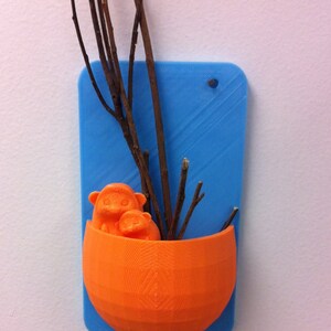 May include: An orange, 3D-printed wall planter with a blue backing. The planter is shaped like a half-sphere and holds two small orange figures and several brown twigs. The blue backing has a hole for hanging.