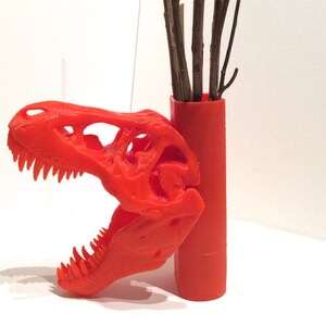 May include: A vibrant orange, 3D-printed Tyrannosaurus Rex skull with sharp teeth, next to a matching orange cylindrical vase holding brown twigs. The skull is detailed and the vase is smooth.