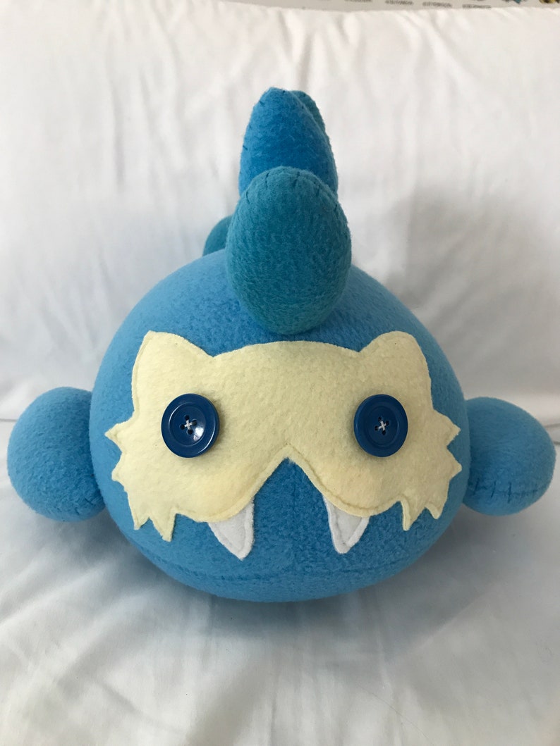 May include: Blue plush toy with a yellow mask and two blue buttons for eyes. The toy has a spiky blue tail.
