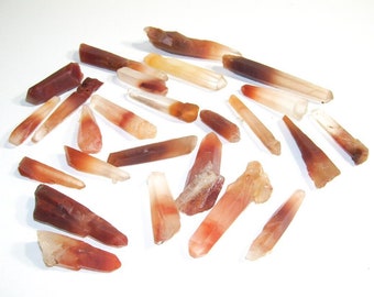 May include: A collection of approximately 20 raw, pointed, red and clear quartz crystals. The crystals are various sizes and shapes, with some having a reddish-brown color at the tip.