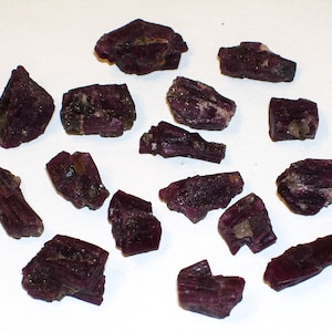 May include: A collection of 15 small, rough, purple-red crystals. The crystals are various shapes and sizes, with some being long and thin, while others are more rounded.