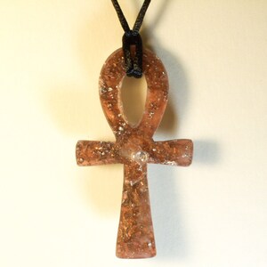 May include: A gold-toned Ankh pendant with a black cord. The pendant is made of resin and has a glittery finish.