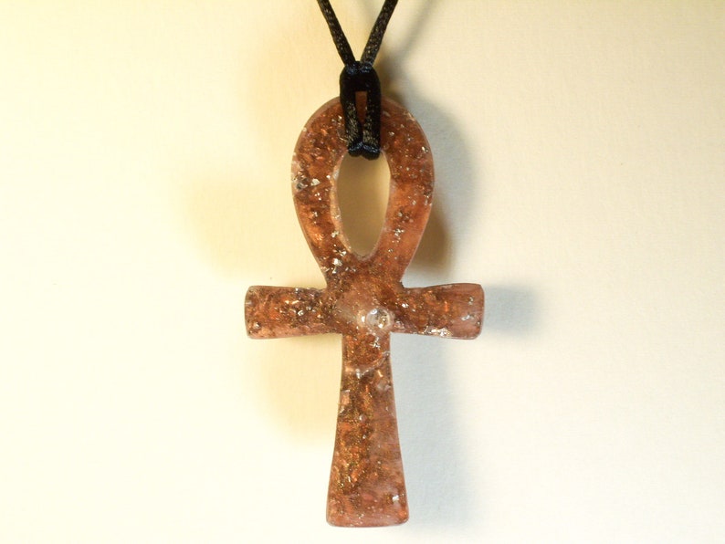 May include: A gold-toned Ankh pendant with a black cord. The pendant is made of resin and has a glittery finish.