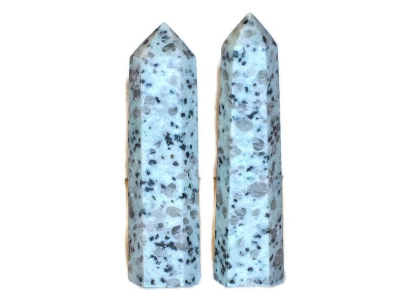 May include: Two light blue and gray speckled crystal obelisks. The obelisks are tall, with a pointed top and a faceted, rectangular body. The crystals have a speckled pattern of gray and black throughout.