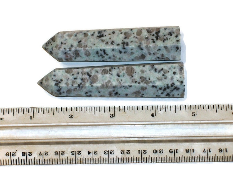 May include: Two polished, light green and brown speckled gemstone obelisks. Each obelisk has a pointed top and a rectangular body. A ruler with inch measurements is in the foreground.