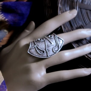 May include: A silver ring with a detailed, abstract design. The ring is worn on a hand, with the design facing the viewer.