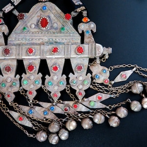 May include: A silver and gold necklace with red, green, and blue gemstones. The necklace has a triangular pendant with a chain of bells.
