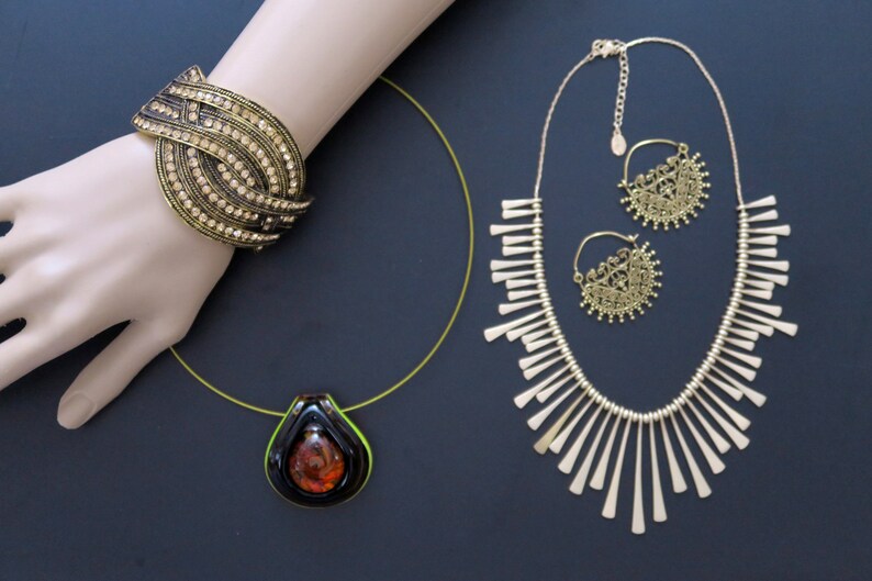 May include: A set of gold-tone jewelry featuring a necklace with a fringe design, a pair of earrings with a circular filigree design, and a bracelet with a multi-layered design with rhinestones.
