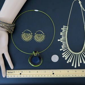 May include: A set of gold-toned jewelry featuring a necklace with a pendant, a pair of hoop earrings, and a bracelet. The pendant is a teardrop shape with a dark blue and green glass design. The necklace is a thin, green wire. The earrings are delicate and have a floral design. The bracelet is wide and has a textured design.