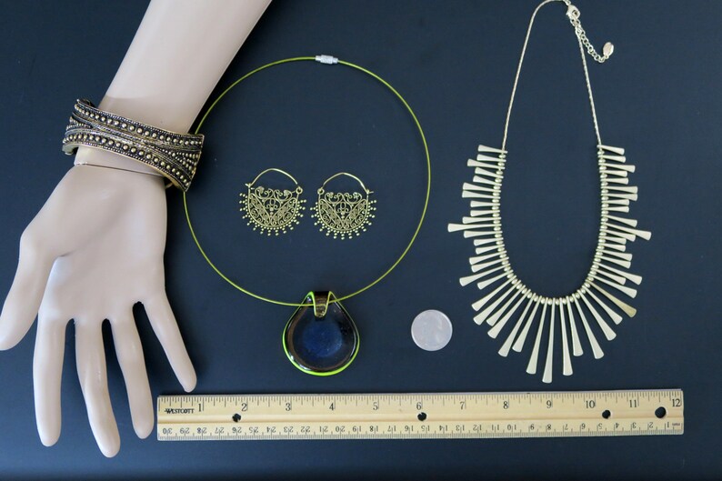 May include: A set of gold-toned jewelry featuring a necklace with a pendant, a pair of hoop earrings, and a bracelet. The pendant is a teardrop shape with a dark blue and green glass design. The necklace is a thin, green wire. The earrings are delicate and have a floral design. The bracelet is wide and has a textured design.