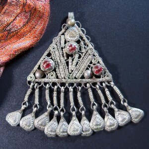 May include: A silver-coloured triangular pendant with red gemstone accents and dangling teardrop charms. The pendant features intricate filigree and beadwork, with a small loop at the top for hanging. A colourful scarf is partially visible in the background.