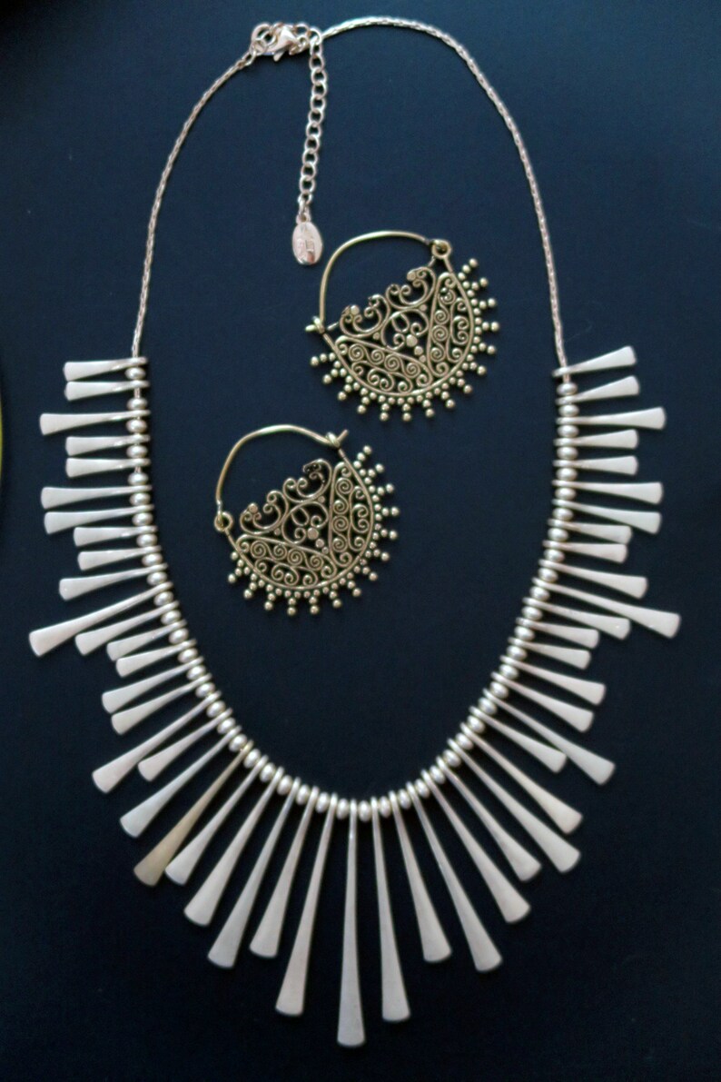 May include: A silver necklace with a fringe design and a pair of gold filigree hoop earrings. The necklace is made of small silver beads and has a delicate, feminine look.