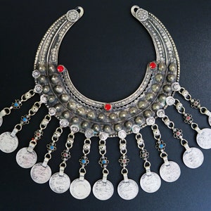Può includere: Una collana d'argento a forma di mezzaluna con monete pendenti. La collana è decorata con pietre preziose rosse e blu.