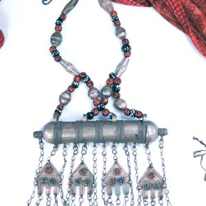 May include: A silver-toned metal necklace featuring a central cylindrical tube pendant with four dangling triangular charms. The charms are inlaid with small red, blue, and green stones. The necklace chain consists of round beads in black, red, and silver.