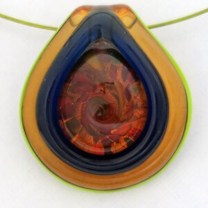 May include: A teardrop-shaped glass pendant necklace with a multi-layered design. The pendant features a swirling pattern of orange, red, and gold glass encased in a blue and gold glass frame. The pendant is suspended from a green cord.