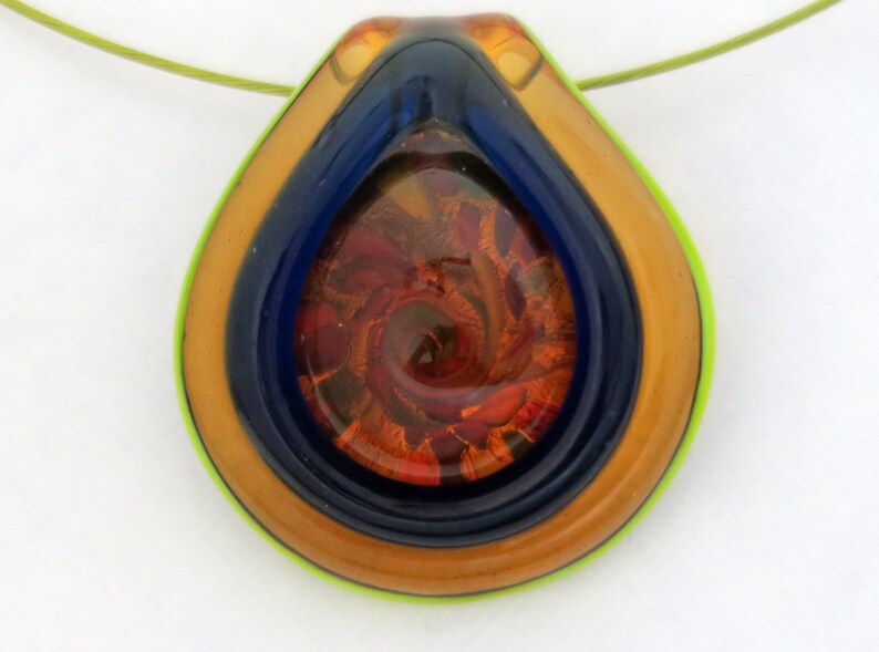 May include: A teardrop-shaped glass pendant necklace with a multi-layered design. The pendant features a swirling pattern of orange, red, and gold glass encased in a blue and gold glass frame. The pendant is suspended from a green cord.