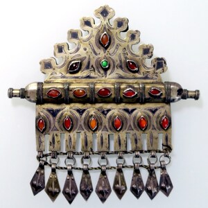 May include: A silver and gold pendant with intricate designs and red and green gemstones. The pendant has a long, narrow shape with a decorative top and a fringe of small, dangling charms.