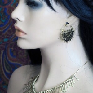 May include: A gold necklace with a fringe design hangs on a mannequin. The necklace is made of small, thin metal pieces that are arranged in a cascading pattern. The mannequin is wearing a pair of gold hoop earrings with a delicate, intricate design.