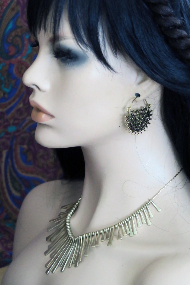 May include: A gold necklace with a fringe design hangs on a mannequin. The necklace is made of small, thin metal pieces that are arranged in a cascading pattern. The mannequin is wearing a pair of gold hoop earrings with a delicate, intricate design.