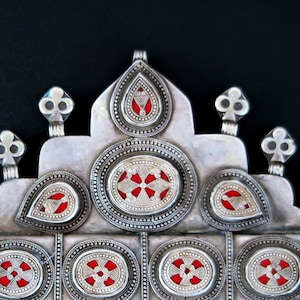May include: A silver pendant with intricate designs and red enamel accents. The pendant features a central oval shape with smaller teardrop shapes and decorative elements.