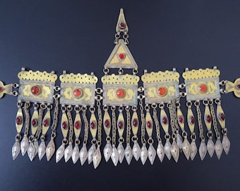 May include: A gold and silver headpiece with red gemstones. The headpiece has a triangular top piece with a smaller triangle hanging from it. The headpiece has a row of rectangular pieces with red gemstones and a row of dangling pieces with red gemstones.