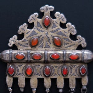 May include: A silver and red jeweled pendant with intricate designs and a crown-like top. The pendant has a long, cylindrical section with multiple red stones and a smaller section with a row of red stones.