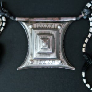 May include: A silver-toned pendant necklace with a geometric design. The pendant features a central square within a larger square, with etched details. The necklace includes black and white beaded strands and black tassels.