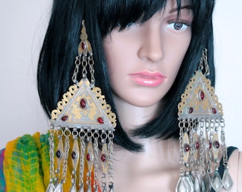 Op de afbeelding: Een paar grote, sierlijke oorbellen met een driehoekig ontwerp. De oorbellen zijn gemaakt van zilver en goud en hebben rode edelstenen. De oorbellen hangen aan de oren van een mannequin met zwart haar.